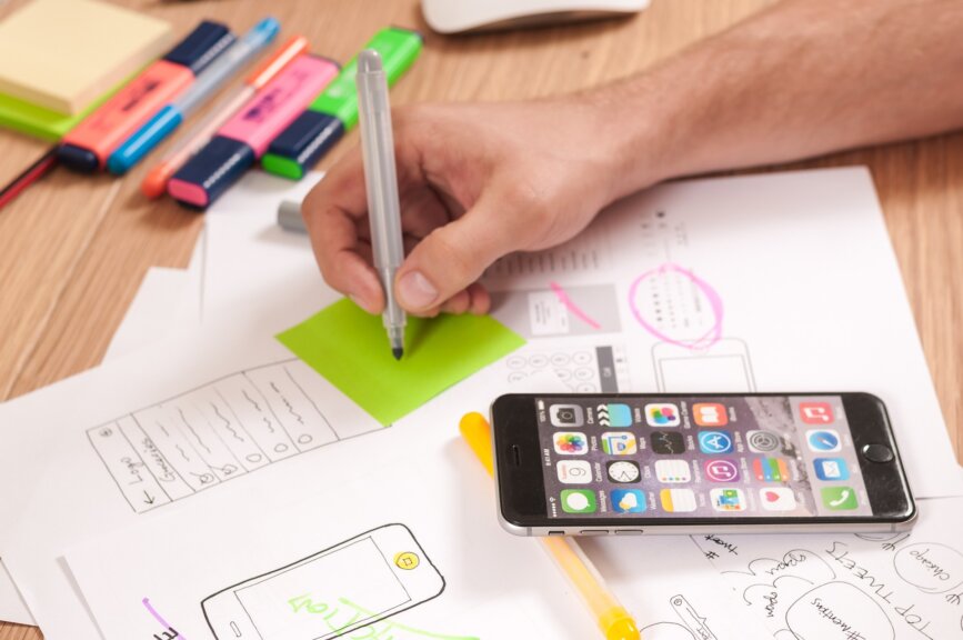 Image of someone's hand hovering over a green post-it note about to write something with lots of paper everywhere underneath, pens and highlighters in the backdrop and a black phone on the homepage screen in the background.