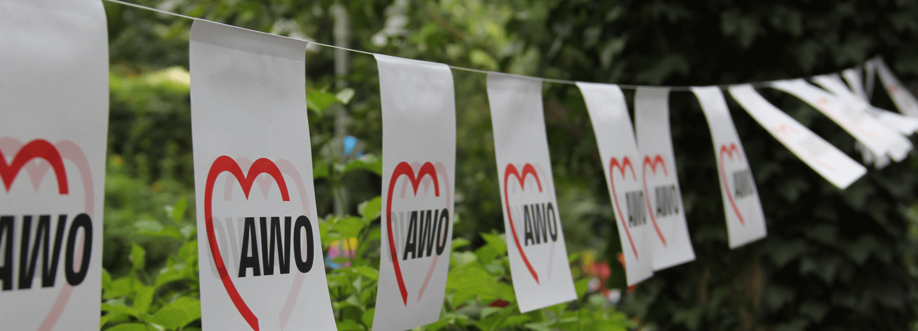 Row of white flags with AWO Berlin-Mitte logo on them wave in the wind with some green shrubbery behind it.